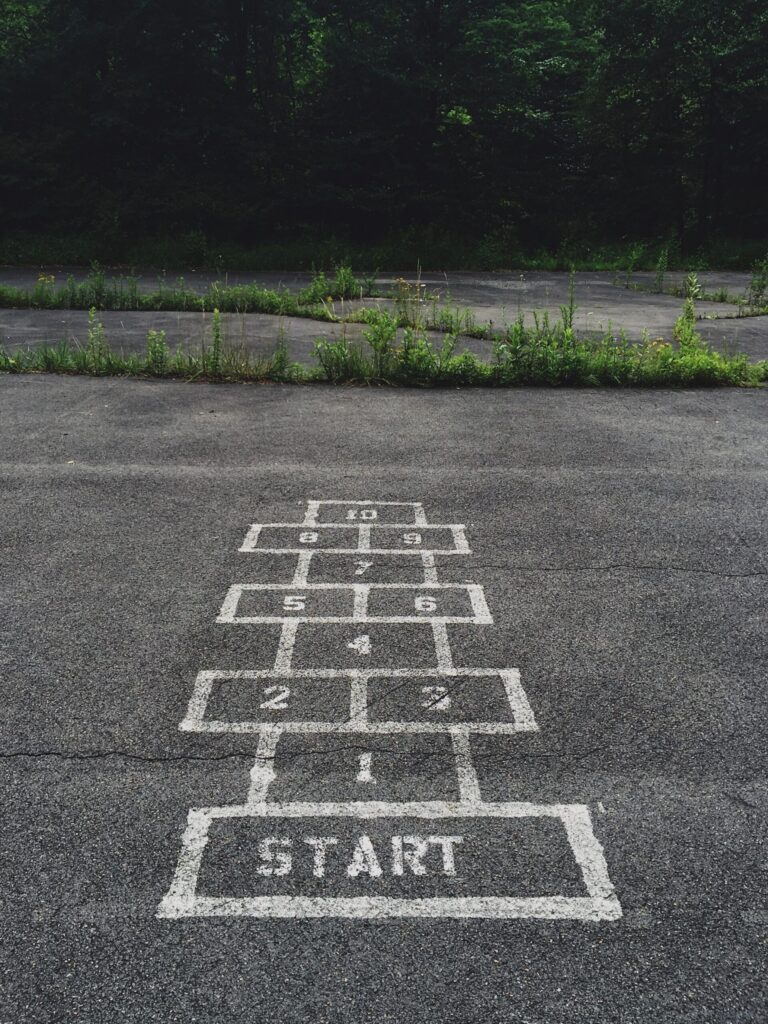 A white hopscotch game is painted on cracked pavement with grass growing through the breaks. The worn surface suggests playfulness, imperfection, and beginning.